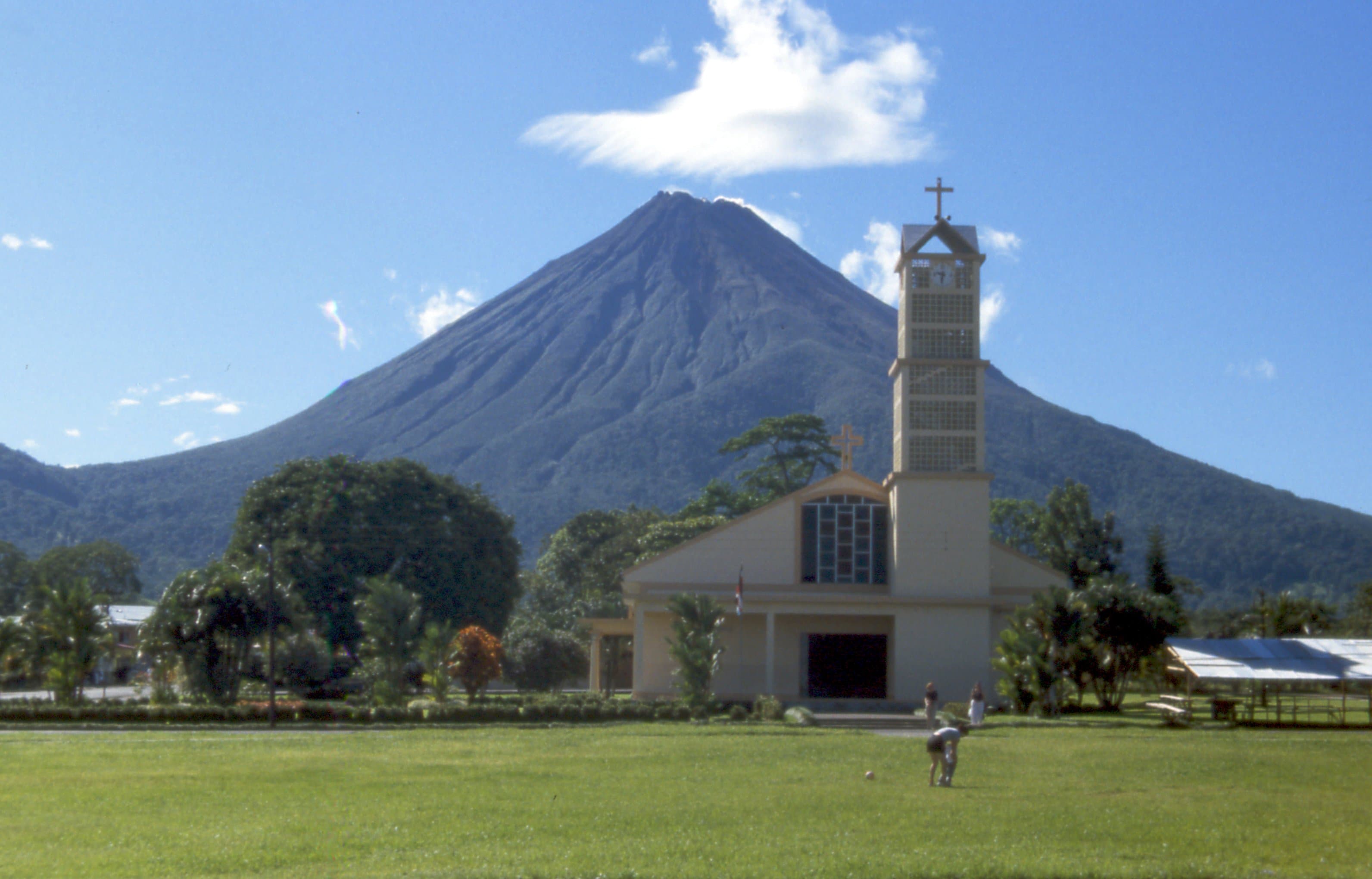 El volcán Arenal detrás de la iglesia de La Fortuna, con el campo verde adelante